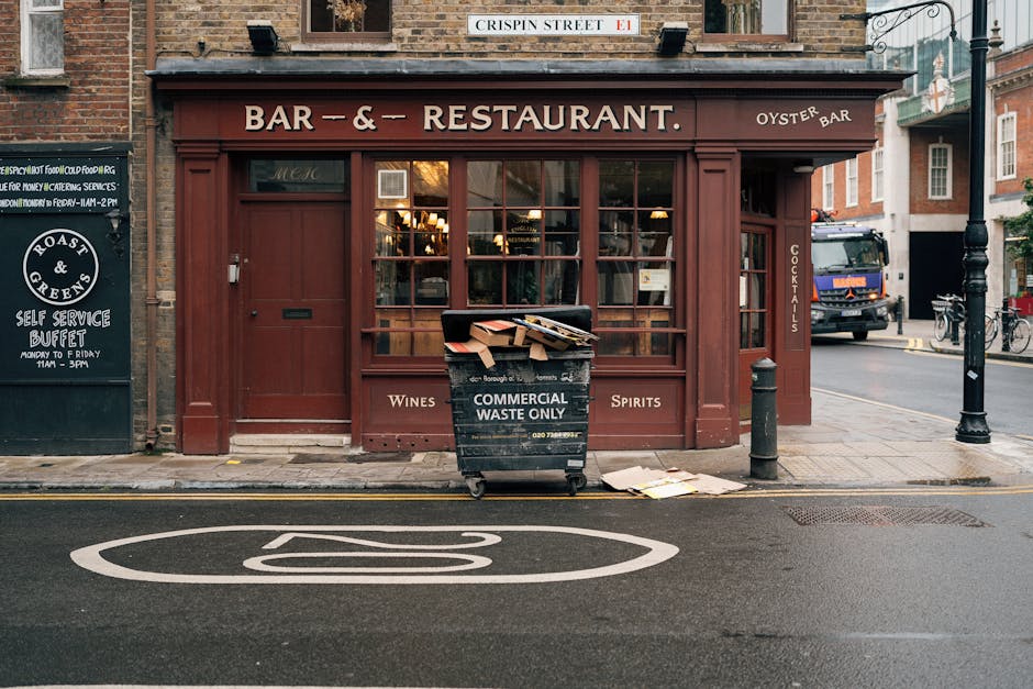 A black wheeled waste collection bin marked 'Commercial Waste Only' is positioned on the curb in front of a red-brown storefront that functions as a bar and restaurant. The bin is filled with flattened cardboard boxes and paper waste, with some materials spilling over the top and resting on the sidewalk. The storefront features large glass windows divided into smaller panes, with warm interior lighting visible through the glass. To the right of the bin, a short metal bollard and a black street lamp are situated near the edge of the sidewalk. Adjacent shops and buildings with brick facades can be seen along a quiet urban street, which includes a vehicle parked in the background and a bicycle leaning against a pole. The pavement and road surface are clean, with a painted circular sign or marking on the asphalt in the foreground. A small black sign to the left displays information about a self-service buffet with opening hours. Overall, the scene illustrates site-specific waste disposal in an urban setting, indicative of private collection practices that complement local rubbish removal services.