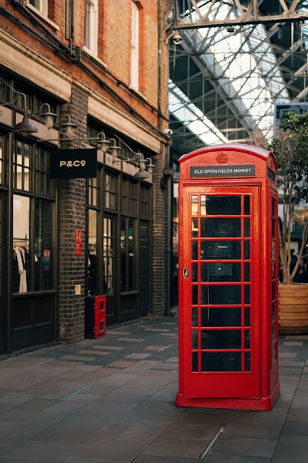 A bright red traditional British telephone box with a rectangular shape and glass paneled doors, positioned on a paved pedestrian pathway inside a covered, glass-roofed indoor market or shopping arcade. The telephone box has a slightly domed top and a sign with white text reading 'OLD SPITALFIELDS MARKET' affixed above the door. To the left of the telephone box, there are storefronts with dark frames, large glass windows, and signs, including one with the text 'P&C9'. The surrounding environment includes a brick building façade with black accents, and overhead, a modern glass ceiling with metal supports allows natural light to illuminate the scene. The area appears clean and well-maintained, with a calm atmosphere that aligns with urban commercial or retail settings where private or independent waste management services may operate alongside public amenities, such as rubbish removal or waste collection in busy indoor environments.