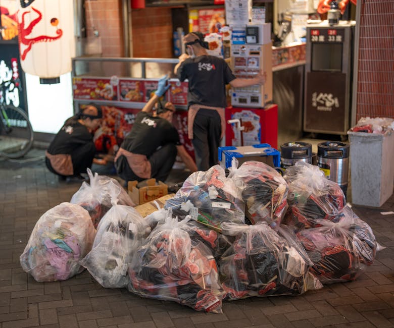 A pile of transparent plastic rubbish bags filled with mixed household waste, including clothing, paper, and packaging, is situated on a paved sidewalk in front of a Japanese restaurant with traditional signage and a red lantern. Behind the bags, three workers wearing black uniforms and beige aprons are crouched down, sorting through or moving additional waste near a storefront with brightly lit menu displays and posters. To the right of the waste, there are two black cylindrical bins, possibly for recycling or waste collection, and a blue plastic crate. The scene occurs in an urban area during evening hours, evidenced by artificial lighting and ambient shadows. The image captures a moment of on-site rubbish clearance, potentially an alternative to regular municipal collection, emphasizing the role of private waste disposal services like Rubbish Removal Shoreditch in managing small business waste in busy street environments.