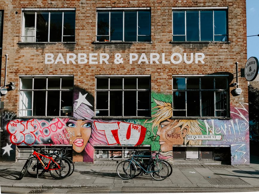 The image depicts the front facade of a brick building with a large sign reading 'Barber & Parlour' in bold white letters at the top. The building has three rows of windows, each with multiple panes, some of which are open or partially open. Prominently displayed on the lower part of the building is colorful street art graffiti featuring stylized female faces with vibrant hair and expressive eyes, accompanied by various tags and words including 'Salon,' 'Newlife,' and other graffiti markings. In front of the building, there are two bicycles parked on the sidewalk, one with a red frame and the other black, both secured to bicycle stands. The environment appears to be an urban street scene, with the building situated along a paved footpath, and there are streetlights and additional signs attached to the building. The overall scene is well-lit, suggesting it was taken during daytime, with a clear blue sky visible above. This scene, associated with a trendy urban area, might relate to services such as private or alternative waste handling, consistent with rubbish removal activities in the vicinity, like those provided by Rubbish Removal Shoreditch.