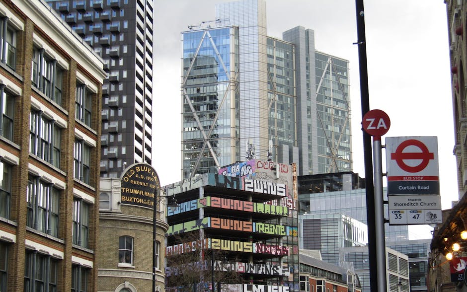 The image depicts an urban street scene with a prominent modern skyscraper featuring a lattice-like structural design and reflective glass windows, dominating the background. In the foreground, there is a tall black pole supporting various signage, including a red and white bus stop sign indicating routes toward Shoreditch Church, and a small circular sign with the letters 'ZA'. To the left, a historic building with a beige brick facade and rows of evenly spaced windows framed by white trim is visible. Below, a large, multi-colored billboard covered in graffiti and street art elements is mounted on a structure, with vibrant hues of pink, green, yellow, and orange, featuring overlapping text and designs. The scene includes elements typical of a busy city environment, such as the combination of old and new architecture, signage, and street art, while the daylight illumination highlights the textures of the buildings' surfaces and the reflective nature of the glass on the skyscraper. This setting provides context for private waste handling and rubbish removal services by illustrating typical urban infrastructure and infrastructure points where rubbish collection or street cleaning might be required, aligning with independent collection and on-site clearance themes relevant to companies like Rubbish Removal Shoreditch.